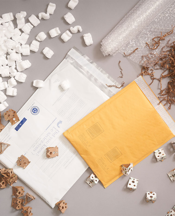 Two shipping envelopes are surrounded by a roll of bubble wrap and piles of different kinds of shipping filler.