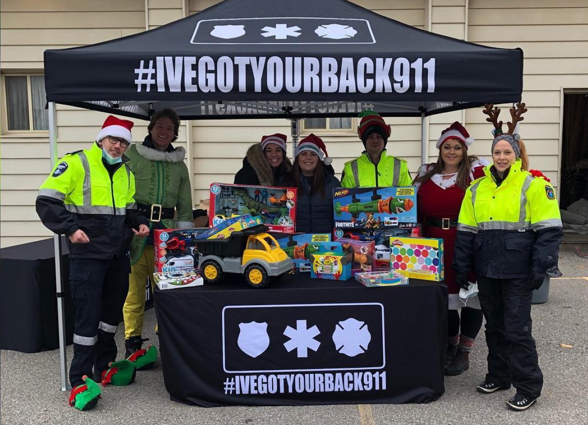 First responders and volunteers in holiday attire at an #IVEGOTYOURBACK911 vendor stand. There are new, unwrapped toys on a table. 