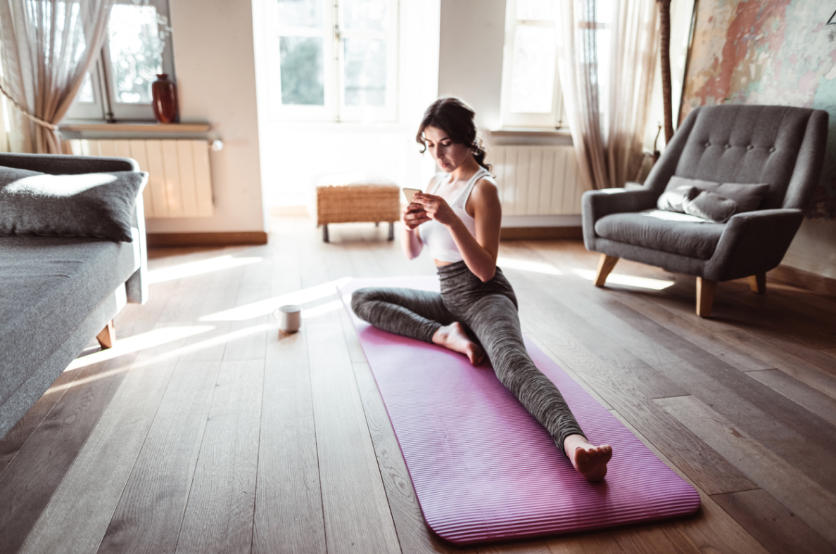 A woman stretches on a yoga mat while looking at her smartphone. 