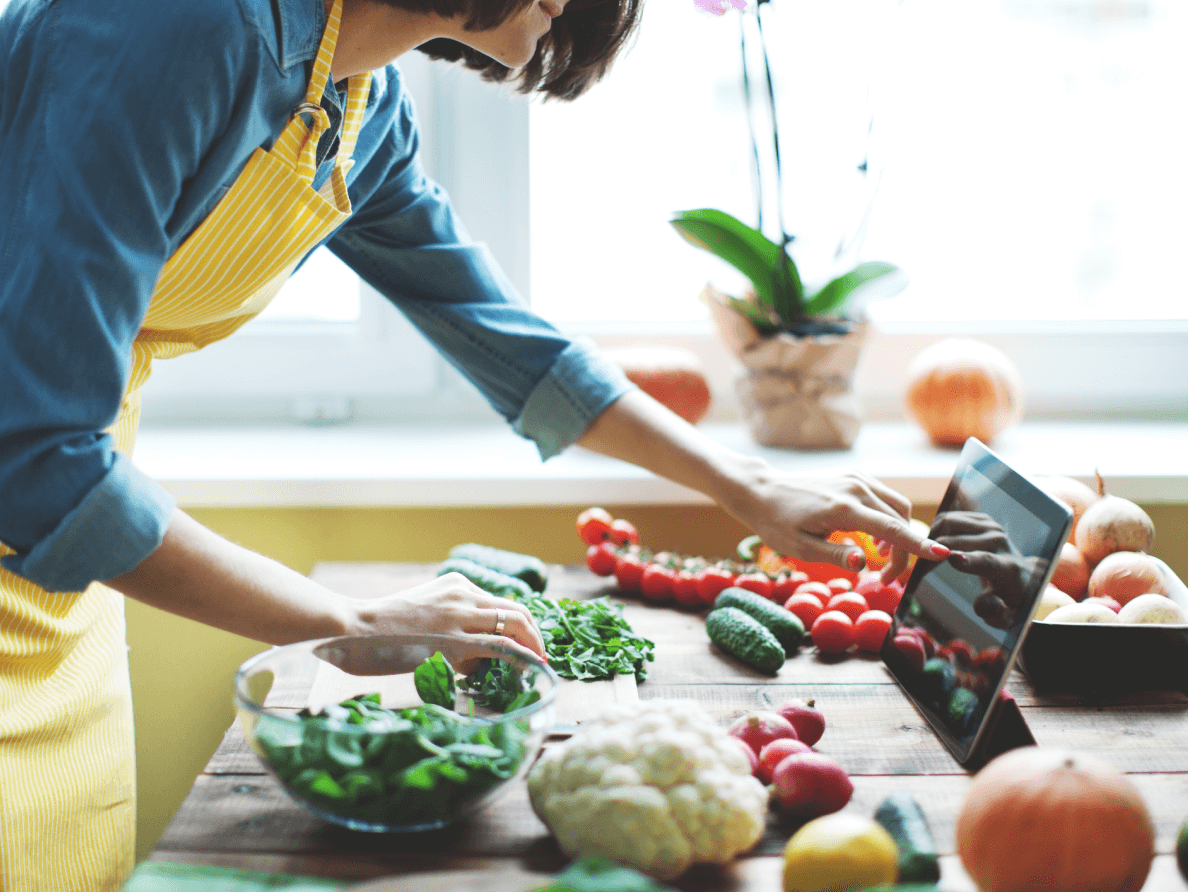 A woman wears an apron. She uses a tablet computer that sits on a table surrounded by fresh vegetables. 