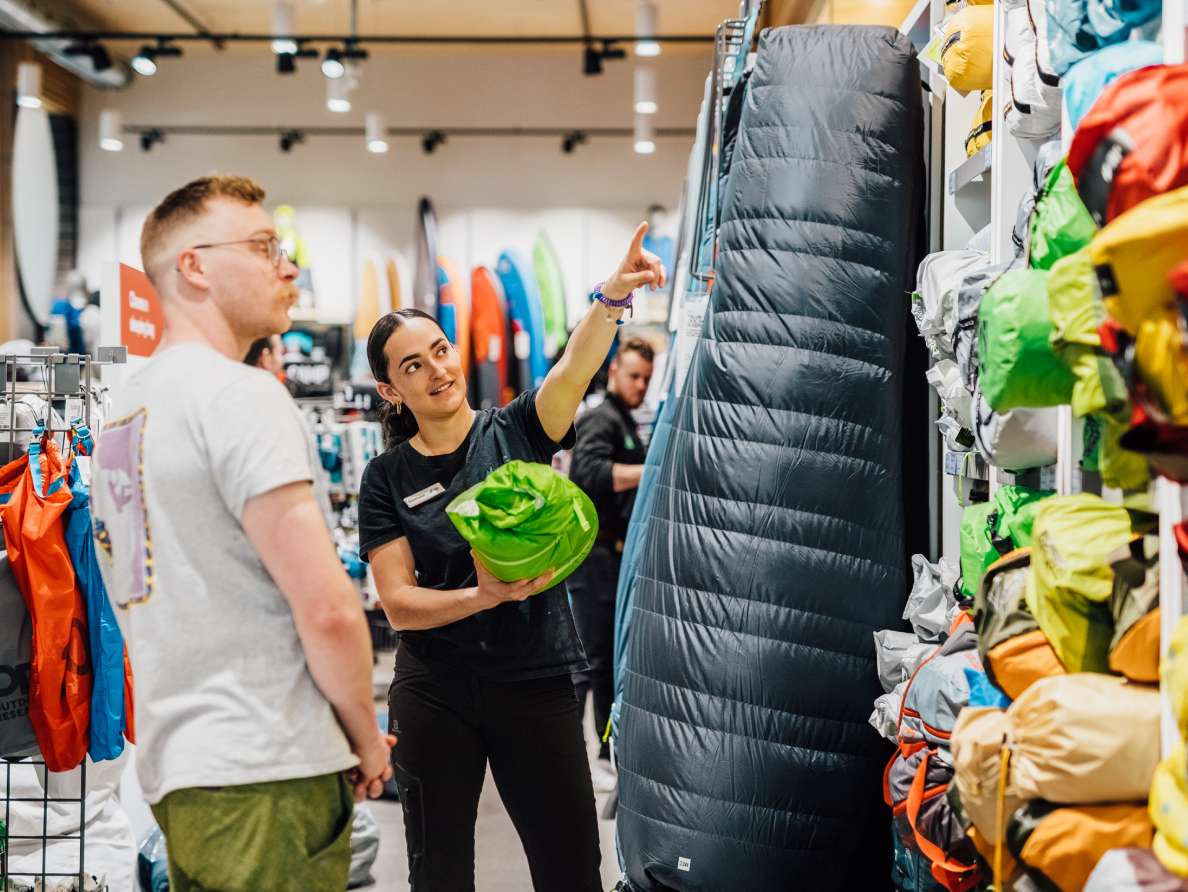 A woman in a black Mountain Equipment Company (MEC) t-shirt points out a sleeping bag to a customer