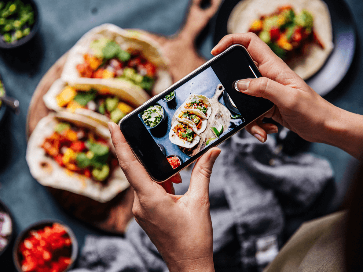 A woman takes a cell phone picture of a table full of food. 