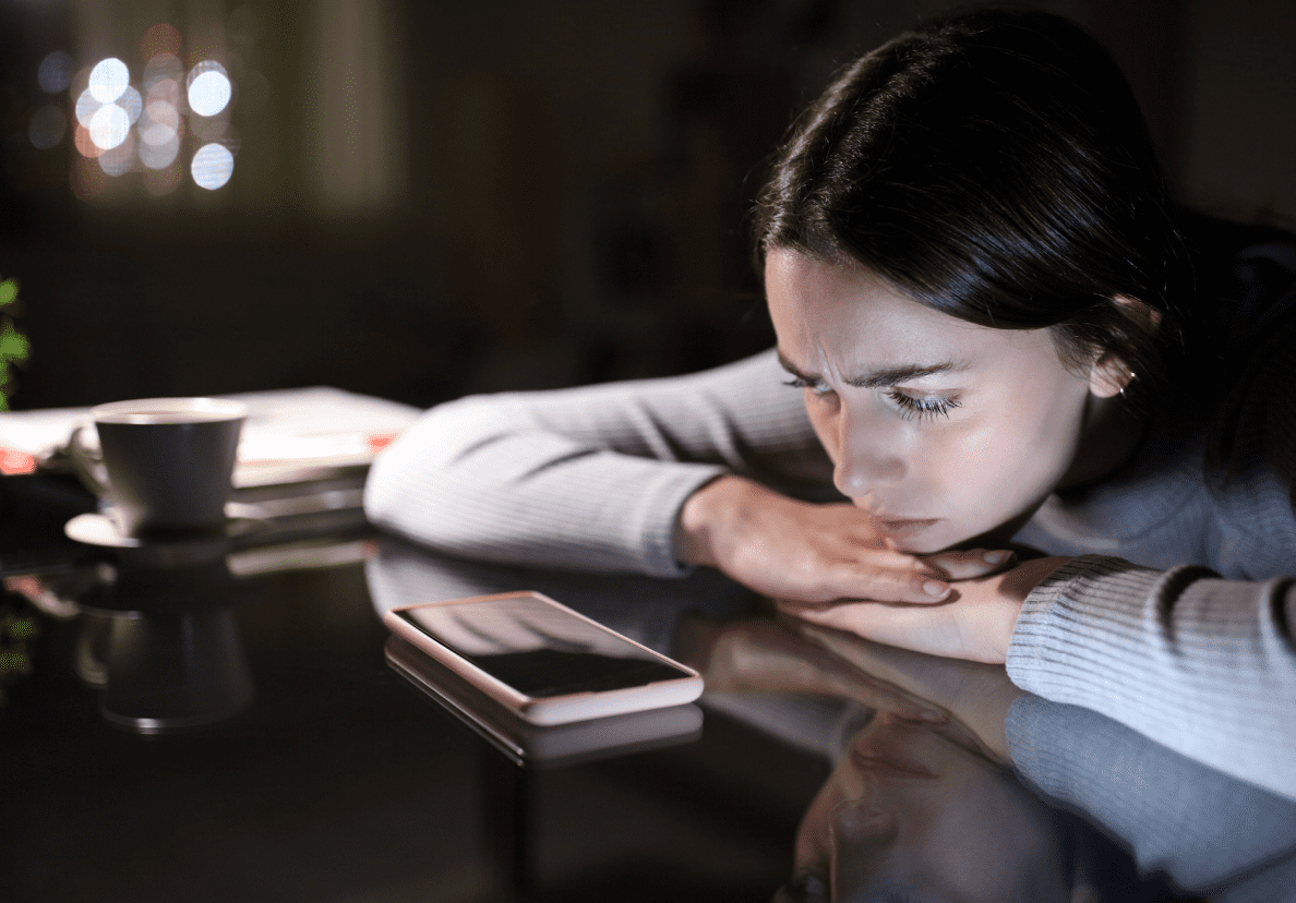 A young woman rests her chin on her hands and stares longingly at silent smartphone with a blank screen. 