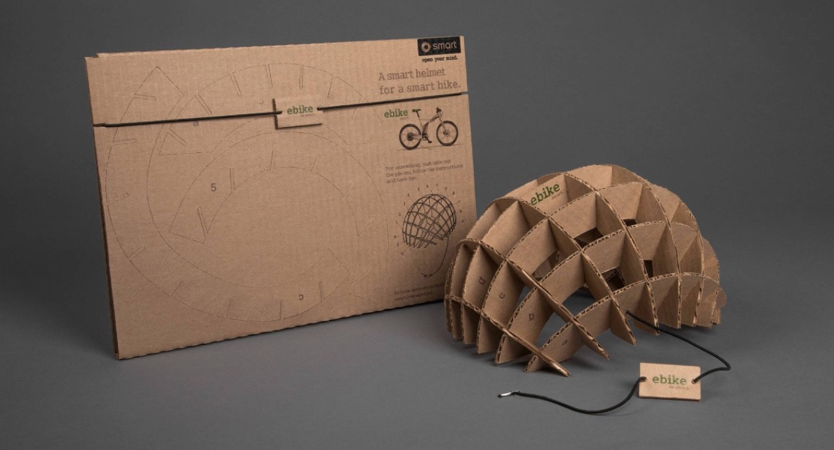 This photo shows off the innovative direct mail campaign from Smart which features a cardboard mail piece that recipients can transform into a cardboard e-bike helmet. An assembled cardboard helmet appears beside the mail piece.