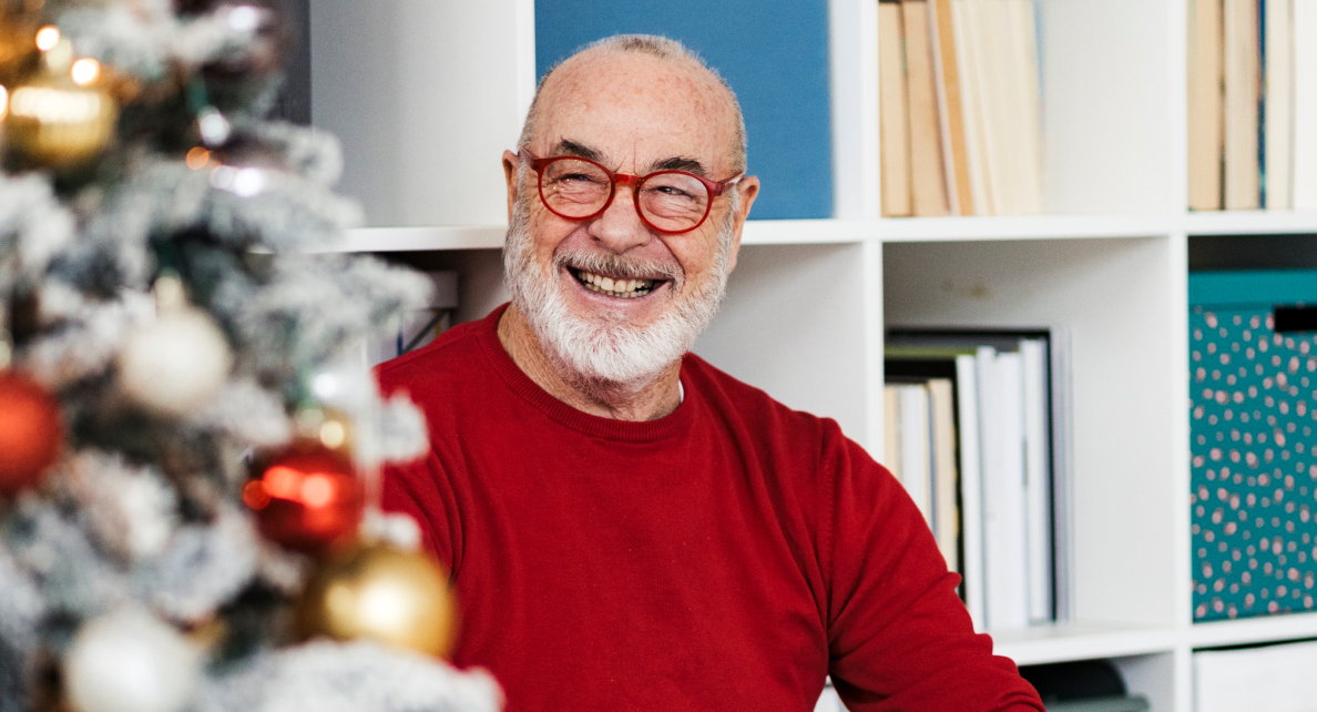 A smiling man with a grey beard sits by a Christmas tree, wearing a red sweater and red glasses.