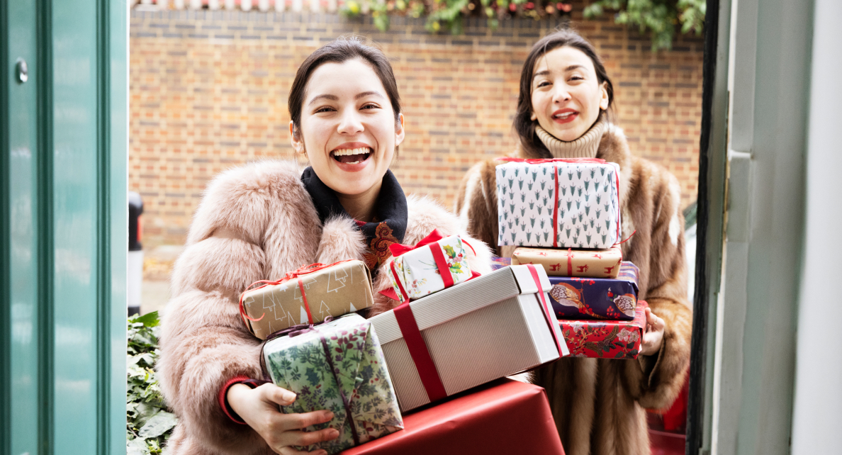 Two smiling women in winter clothes hold several wrapped gifts in their arms. 
