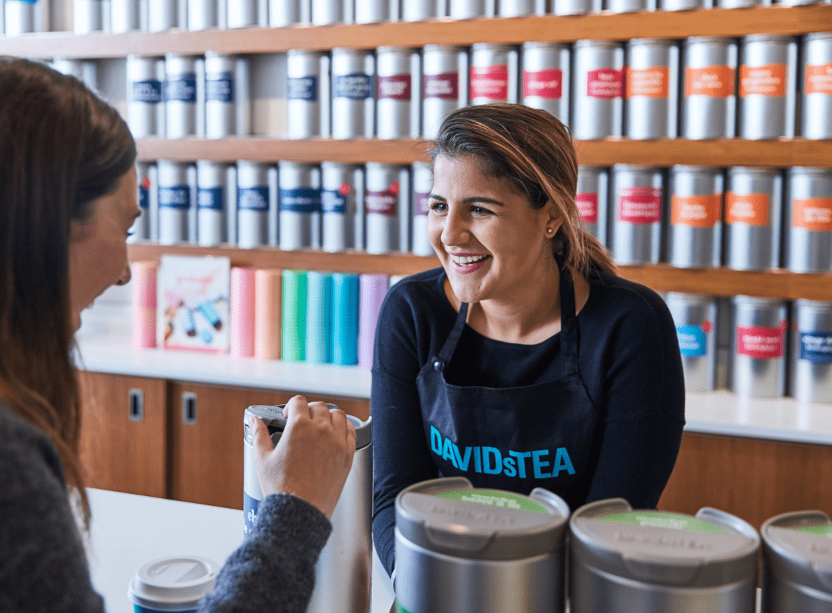 A DAVIDsTEA employee smiles at a customer. She stands in front of a wall of tea canisters.