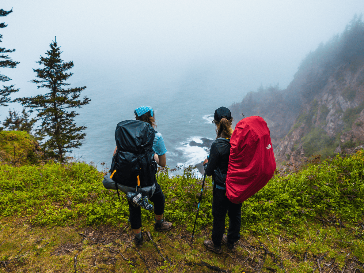 A pair of hikers, wearing large backpacks and carrying hiking poles, stand on a cliff and admire the view of the sea.