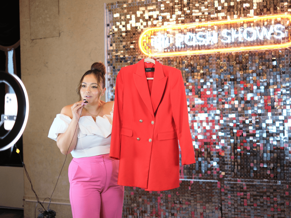 A woman holds up a woman’s red, double-breasted blazer at a Posh Show, live shopping event.