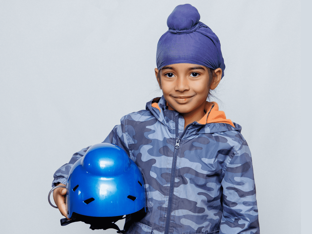 A Sikh child with a top knot head wrap smiles and holds a blue helmet from Bold Helmets.