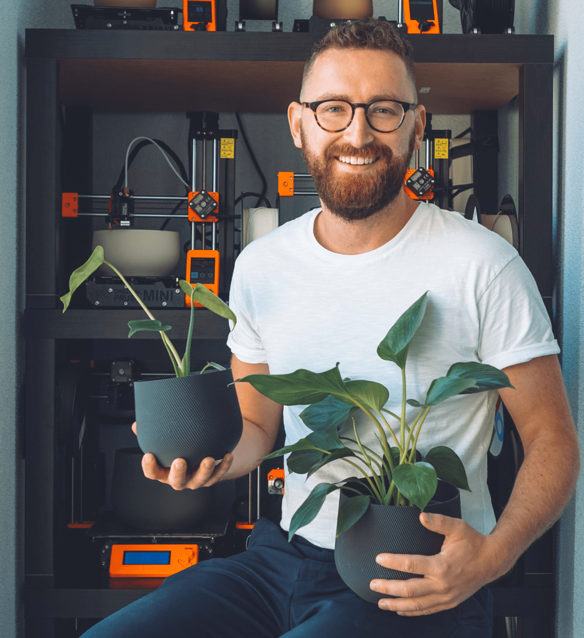  Evan McDougall, founder of Conifer Homewares, holds two dark grey 3D printed plant pots that each have a plant in them. He sits in front of a 3D printer. Evan McDougall, fondateur de Conifer Homewares, tient deux pots à plante gris foncé imprimés en 3D contenant chacun une plante. Il est assis devant une imprimante 3D