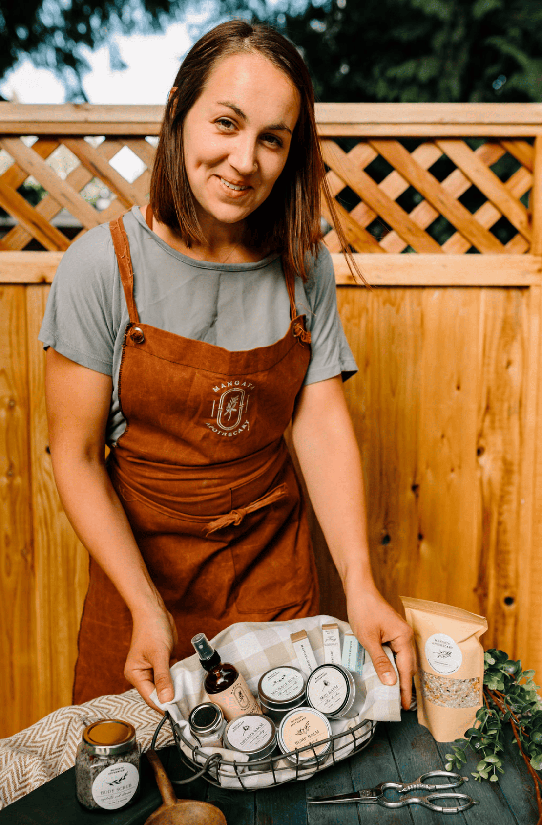 Founder of Mangata Apothecary, Stephanie Henderland, wears an apron and holds a basket of her products.