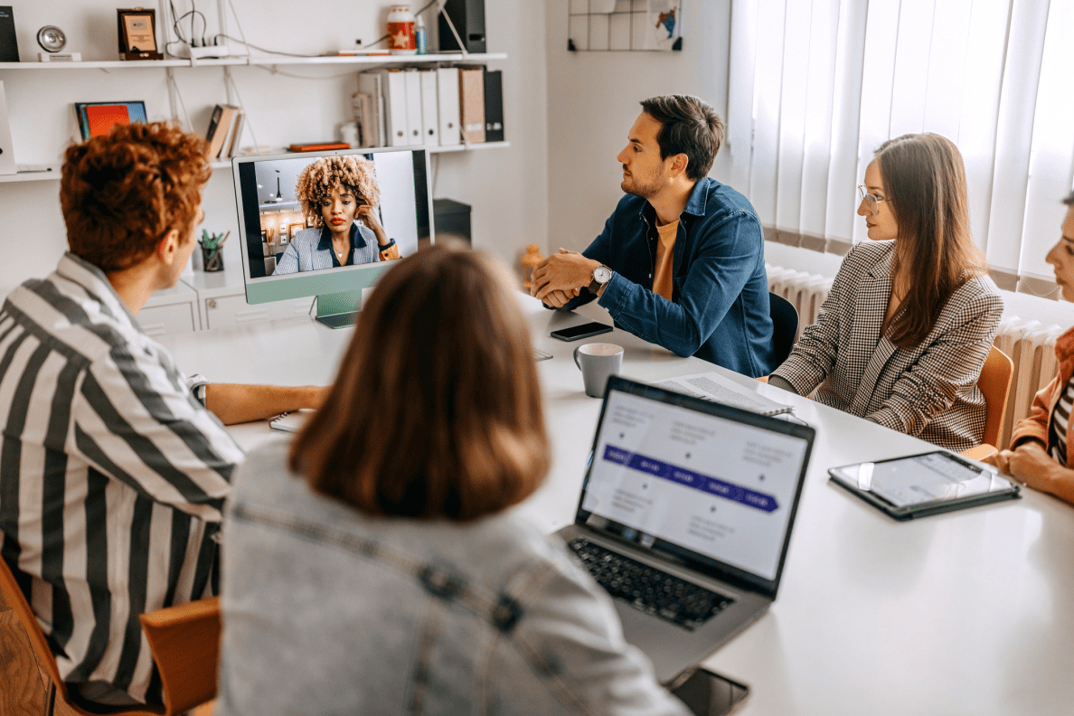 2 men and 2 women sit in a small board room, having a meeting. They share a single video screen. A woman appears virtually on the screen.