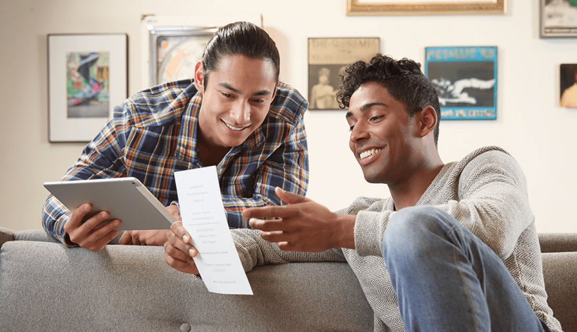 A young man holds a tablet computer as he leans over the back of a couch. Another young man sits on the couch reading a piece of mail.