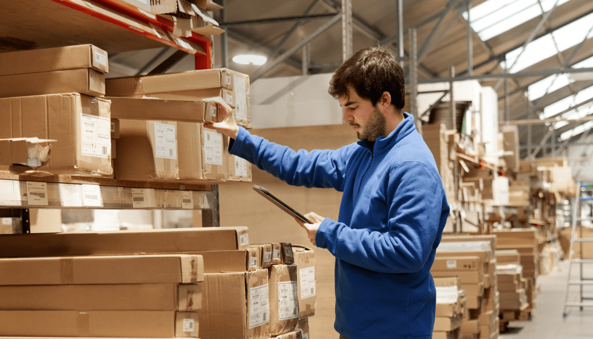 A man stands in a warehouse, looking at a clipboard, as he pulls an inventory item from a large metal shelf.