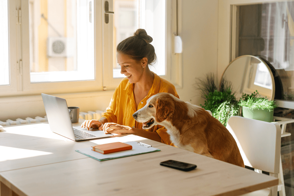 A woman sits at a table in her home. She smiles as she looks at the screen of her laptop. A dog sits in the chair next to her.