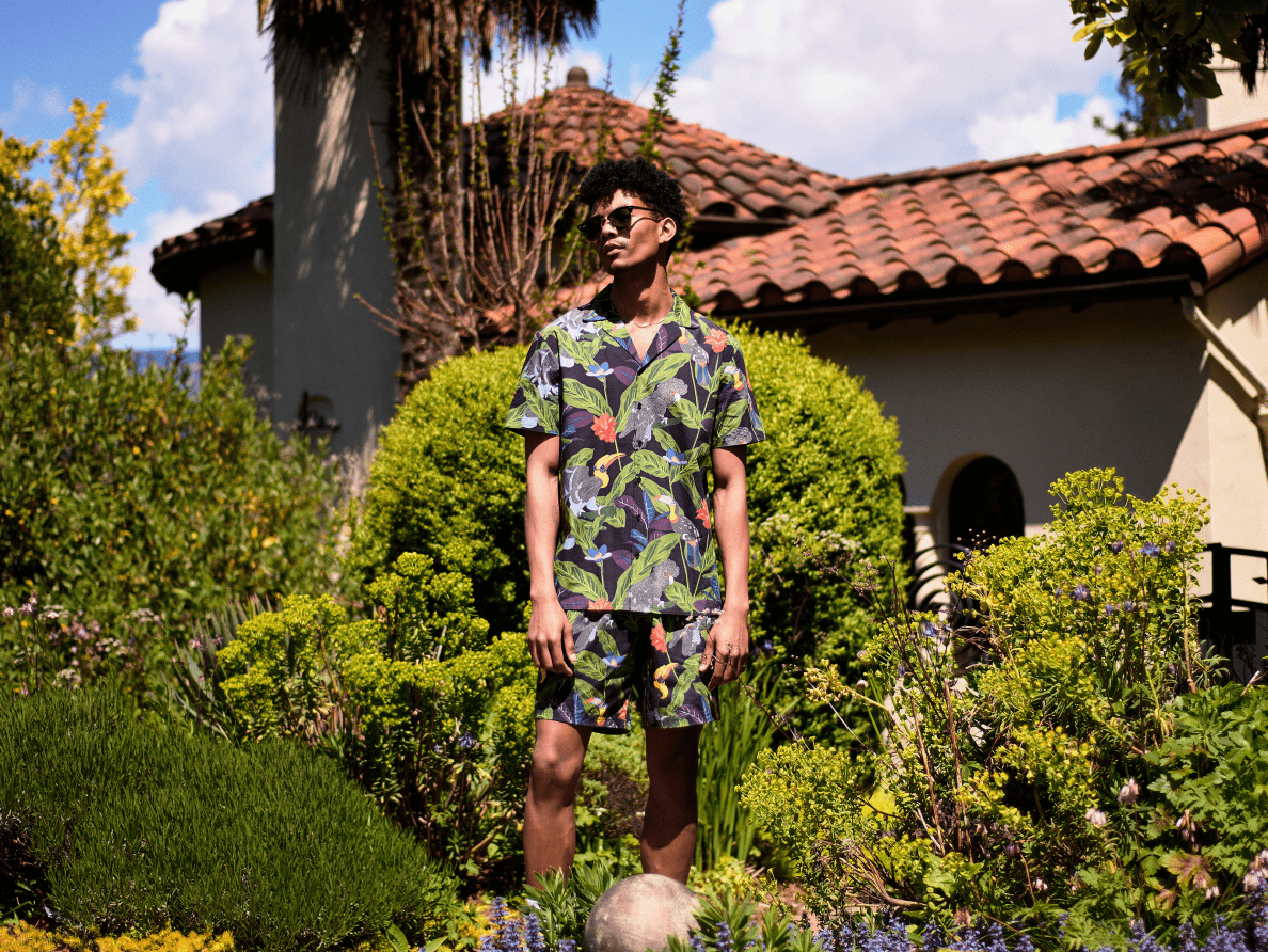 A male model wears a shirt and matching shorts with a tropical pattern from Poplin & Co. and stands in a garden outside a house.