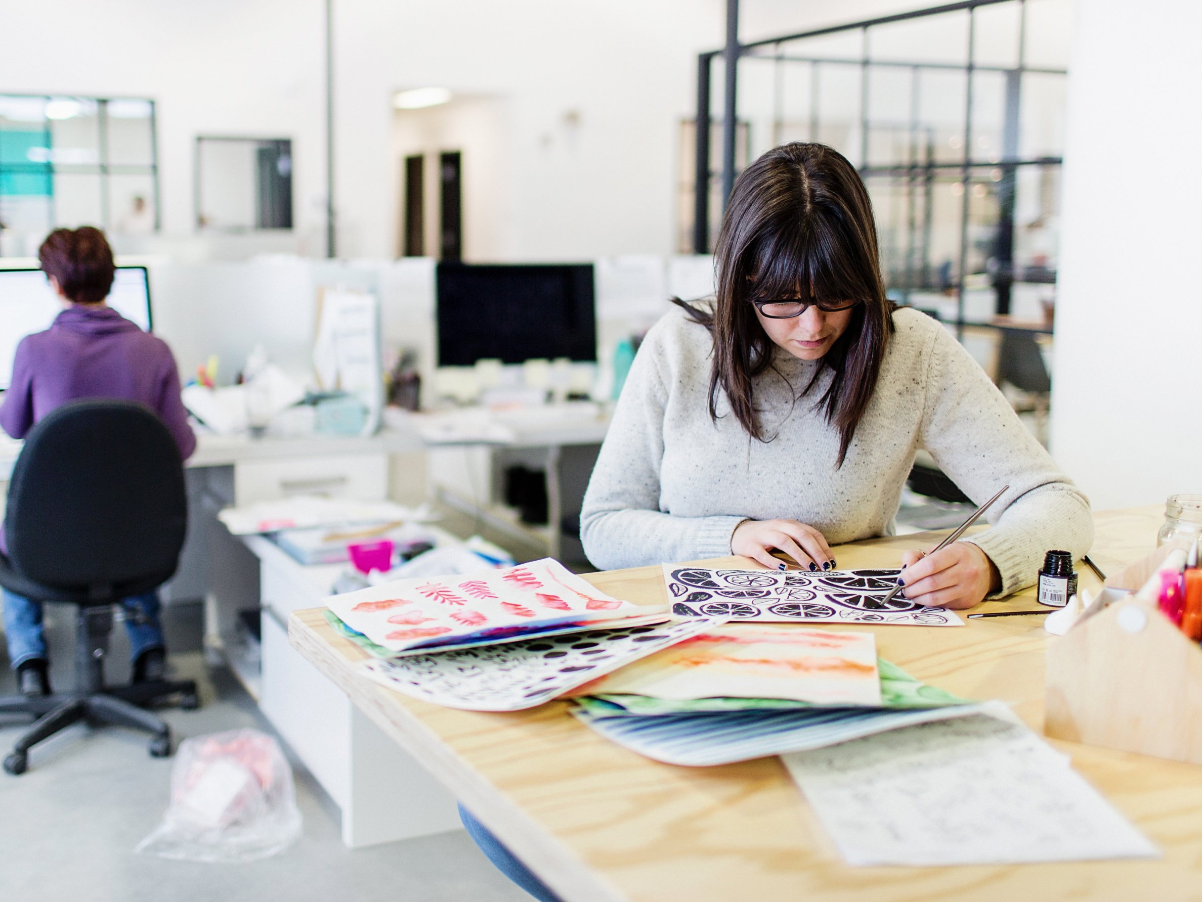 A designer works at a table, painting an image. On the table are several other colourful designs.