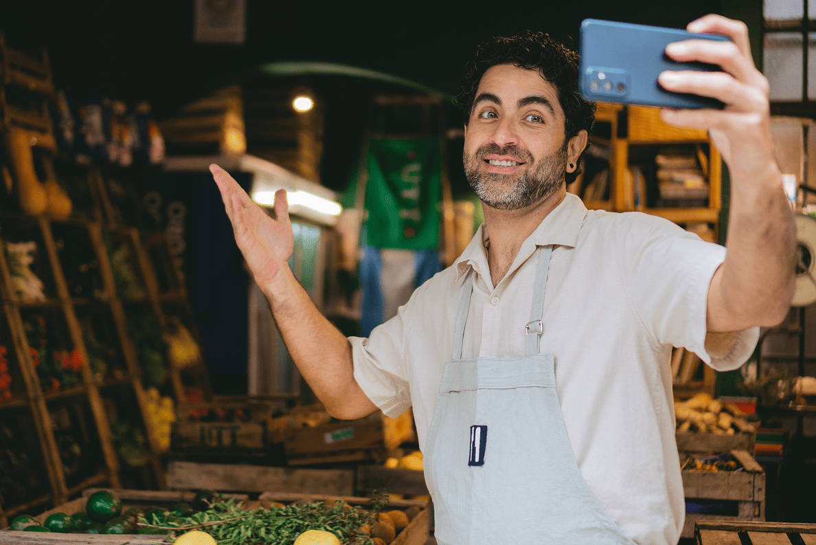 Smiling small business owner wears an apron and takes a selfie in vibrant produce market, amid crates of fresh fruits and vegetables.