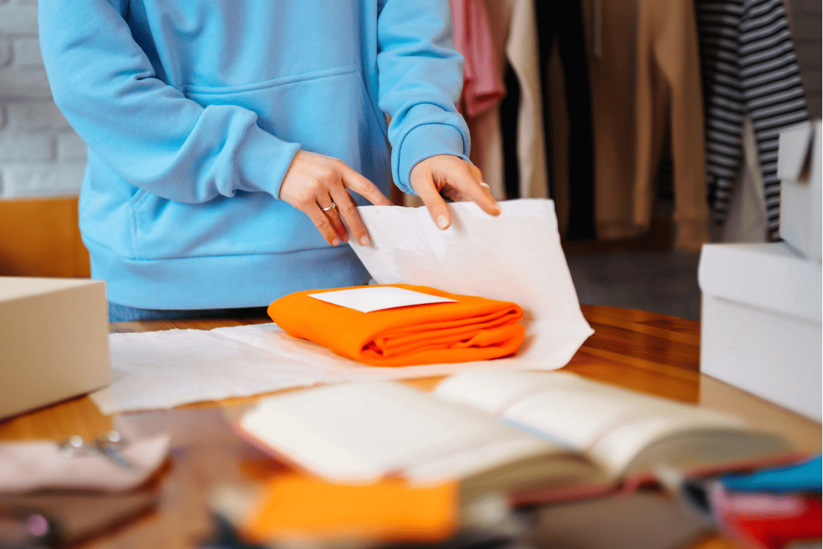 Person in blue hoodie carefully wraps orange clothing in tissue paper at fashion workspace, near hanging garments.
