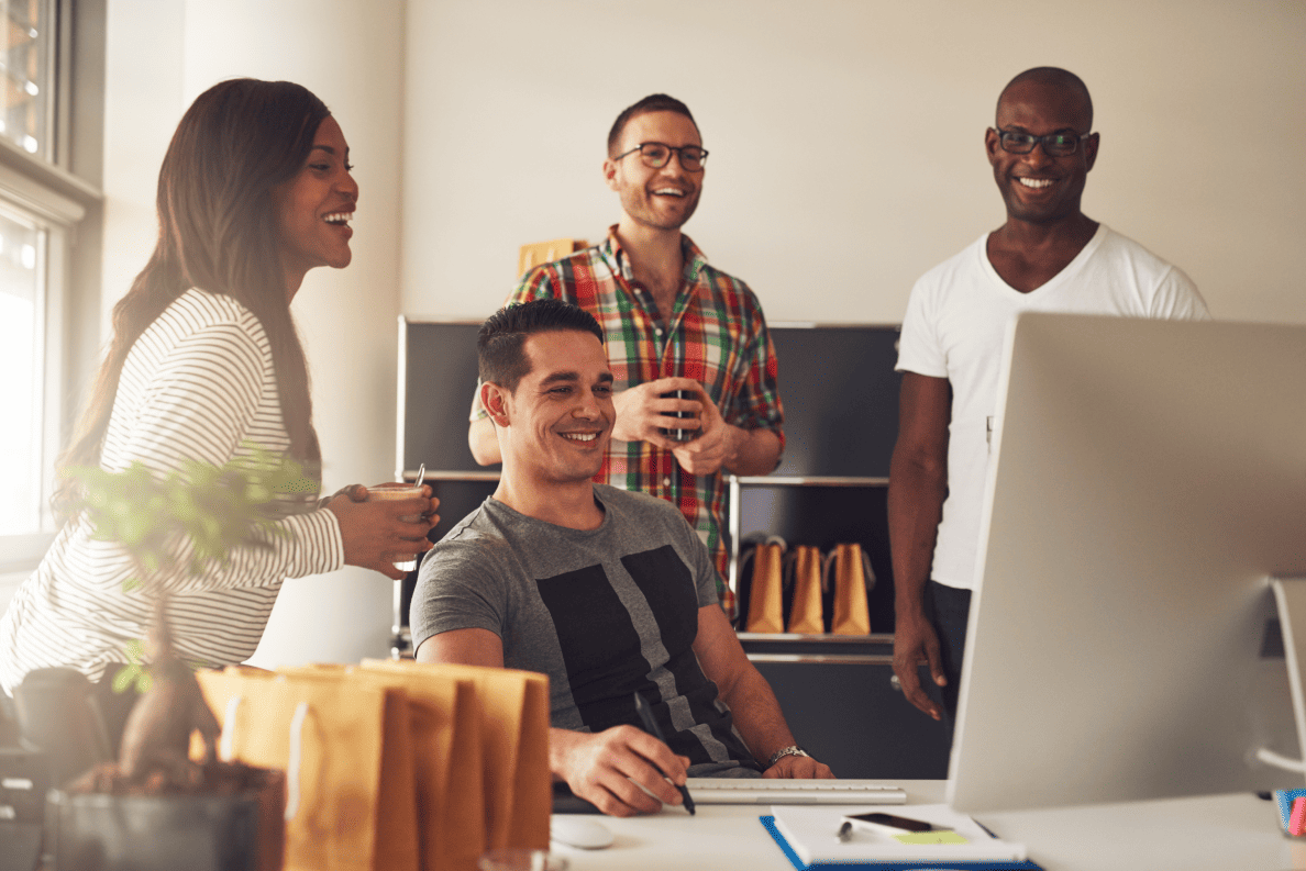 Four casually dressed people gather around a computer in a bright workspace, smiling and collaborating in a relaxed, productive setting.