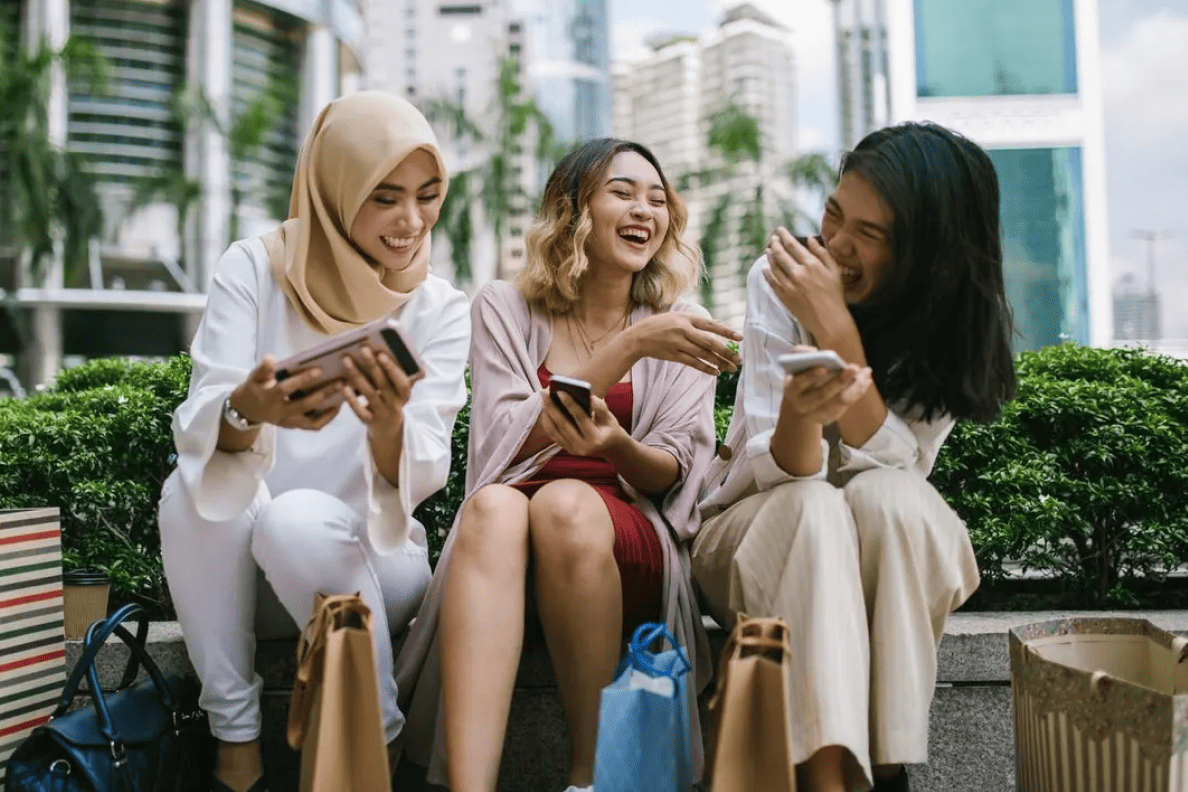 Three young women smile and laugh as they hold smart phones and sit outside in an urban setting.