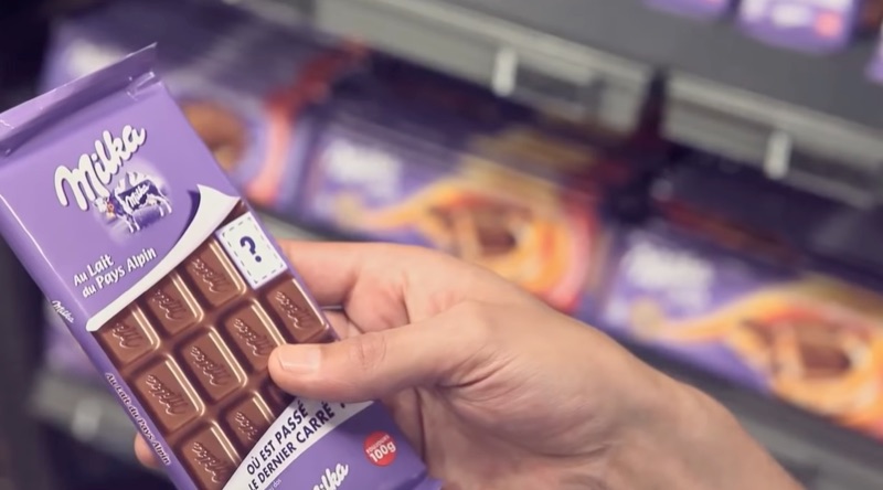 Une tablette de chocolat Milka dans son emballage; un point d’interrogation est imprimé à l’endroit où il manque un morceau. 