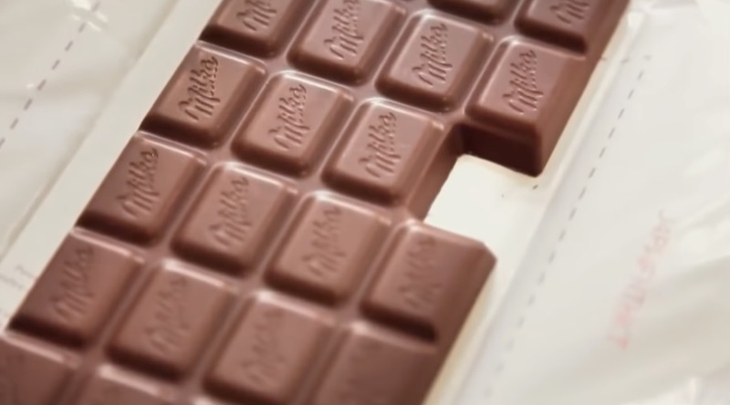 Une tablette de chocolat Milka posée sur son emballage ouvert; il manque un morceau. 