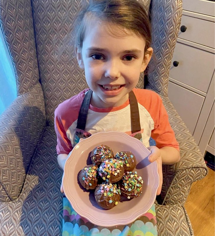 Une petite fille en tablier sourit en présentant un bol de pâtisseries au chocolat avec des vermicelles multicolores.