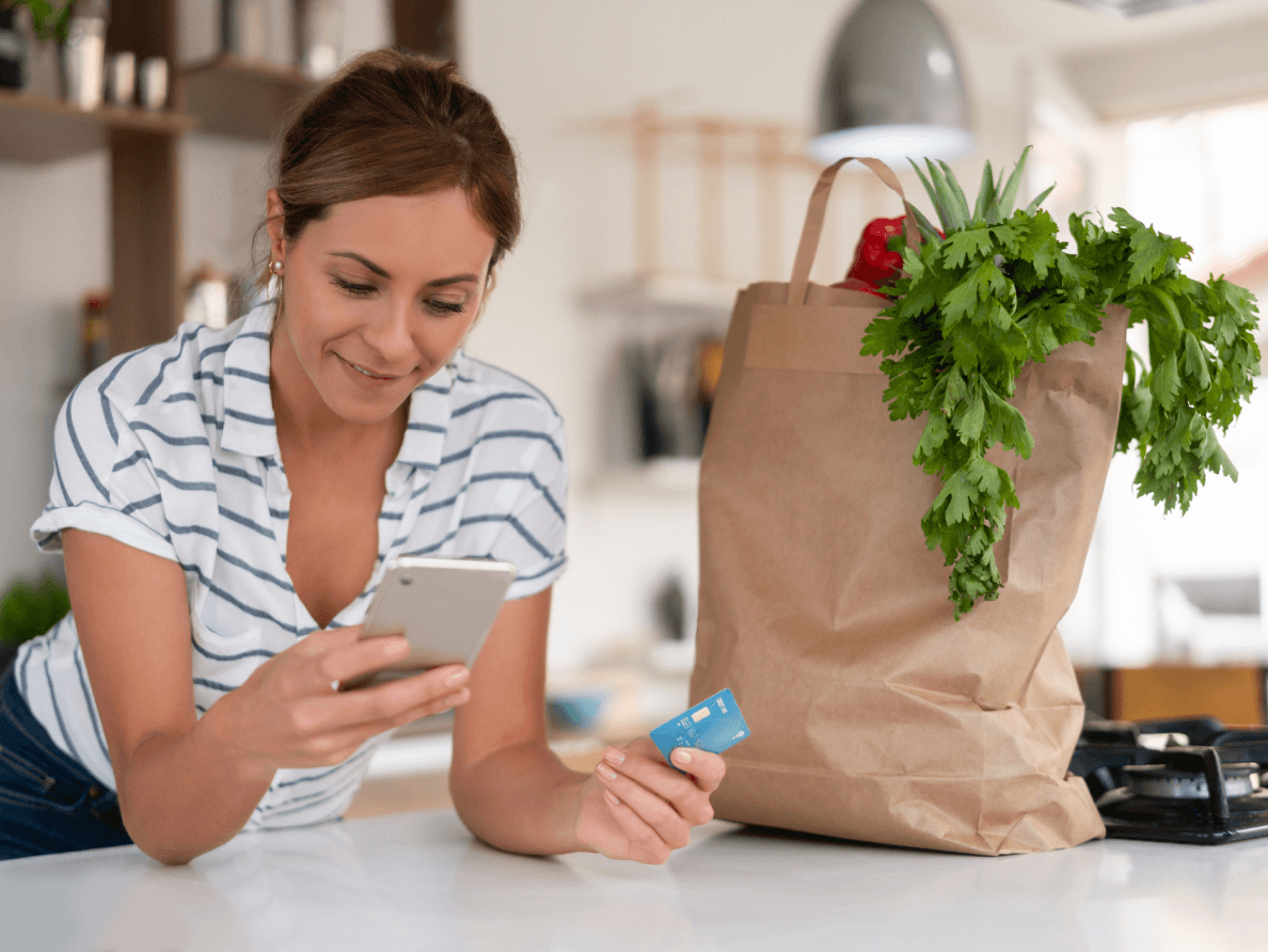 Un sac d’épicerie à ses côtés, une femme consulte son téléphone intelligent, une carte de crédit à la main. 