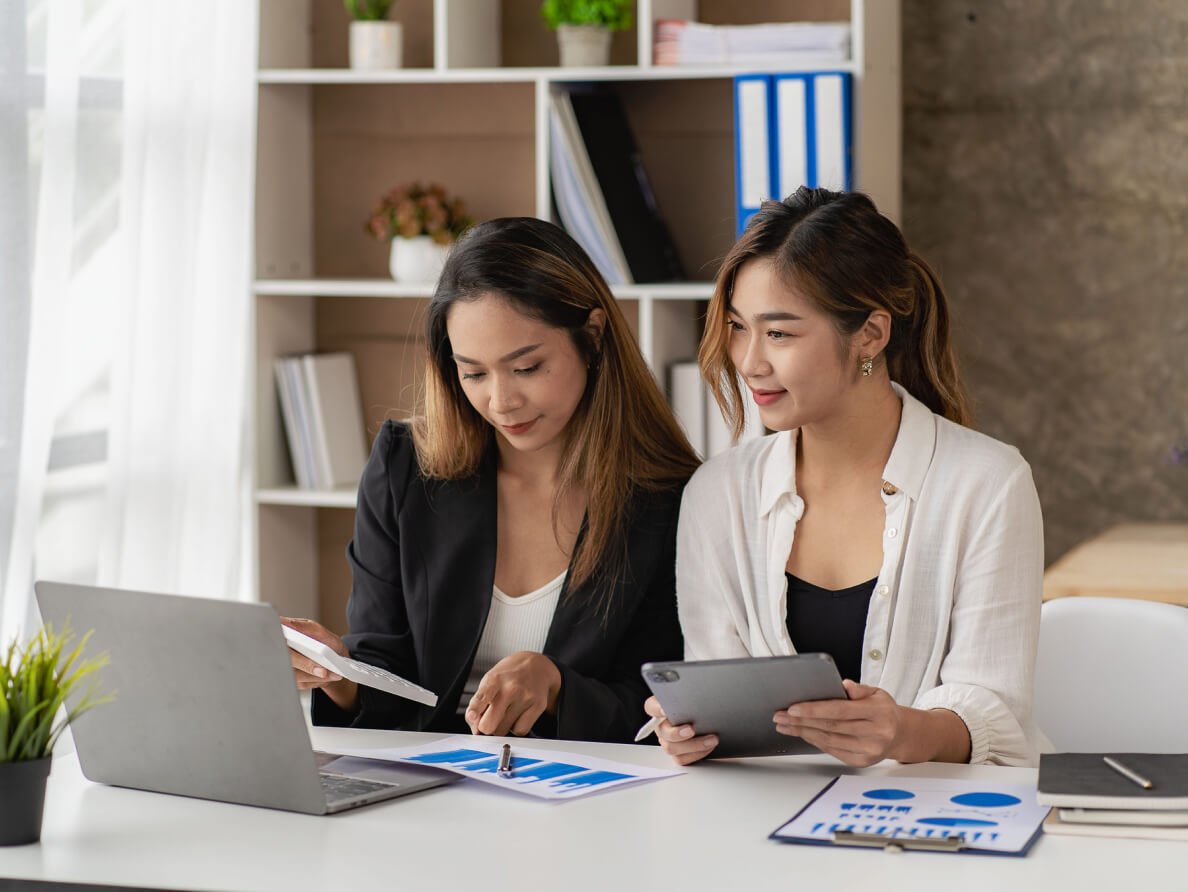 2 femmes assises à une table de travail dont une pointant un graphique sur papier et l’autre regardant l’écran d’un ordinateur portable