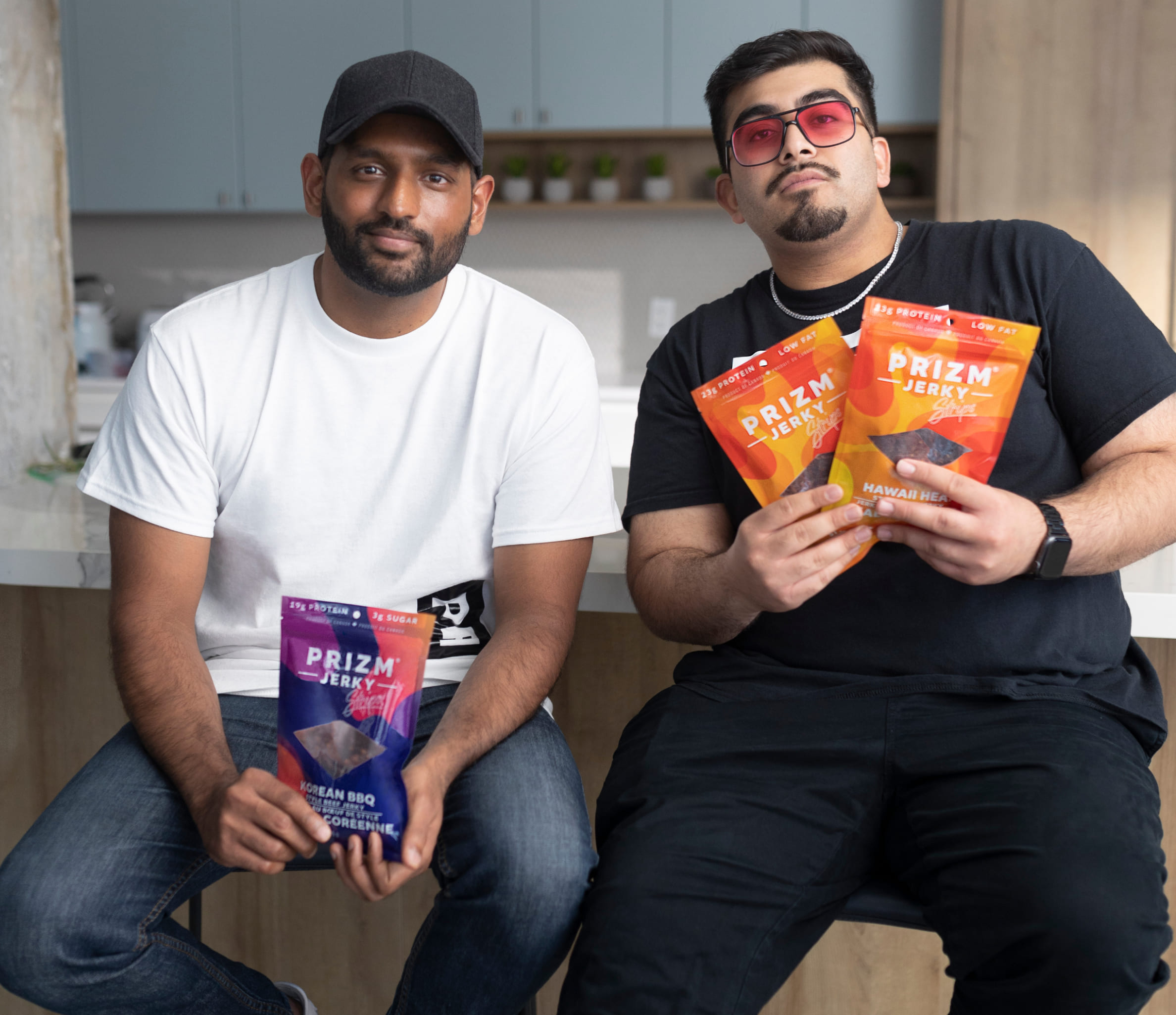 Shaan and Humza of Prizm Foods sit on stools in front of a kitchen island holding purple and orange bags of their jerky.