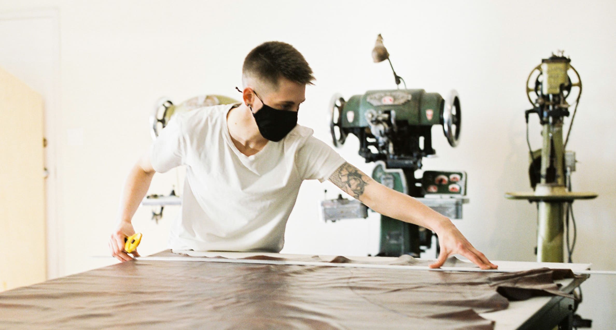 Ariss Grutter, qui porte un t-shirt et un masque en tissu, mesure un tissu sur une table devant deux machines à textile