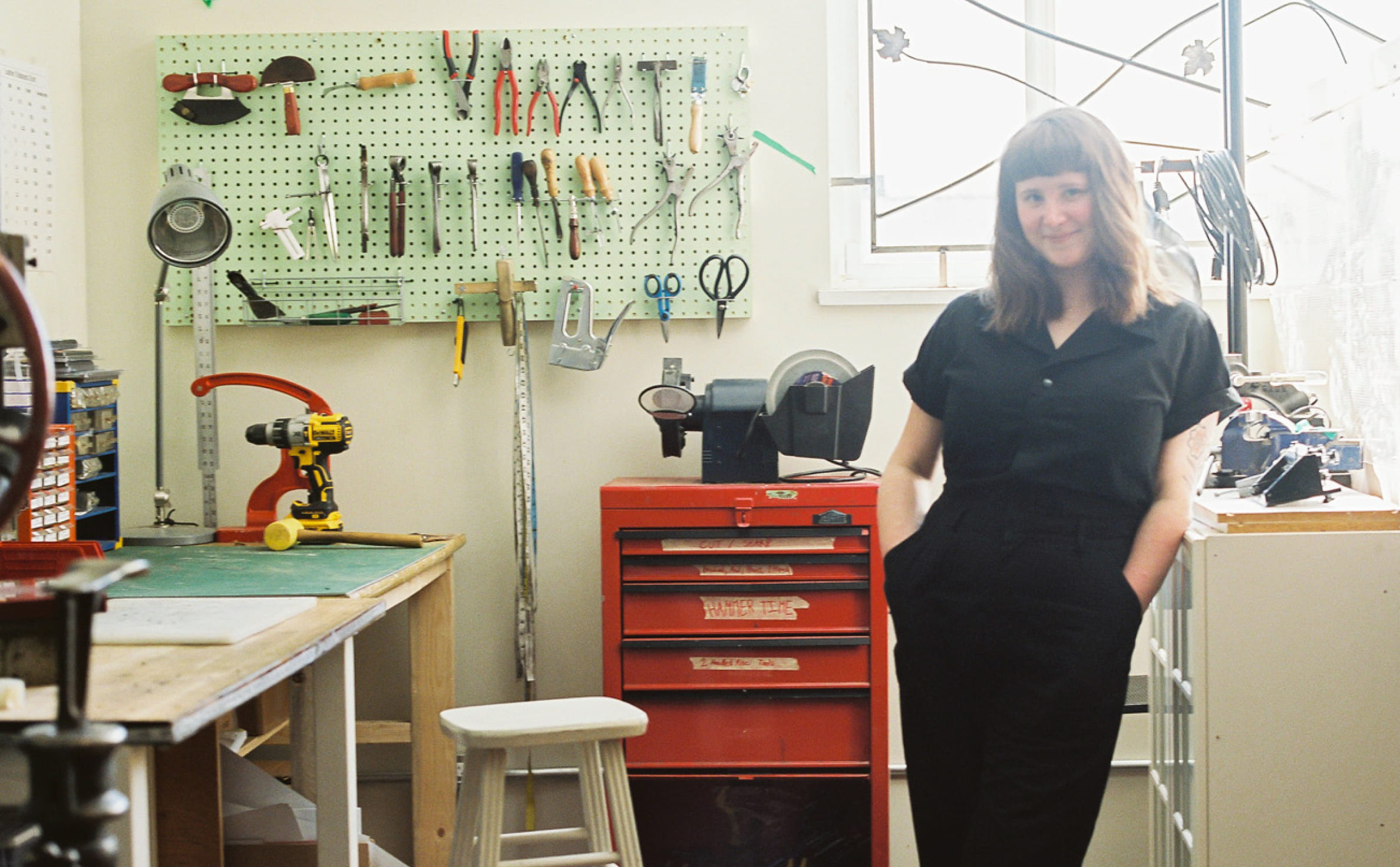 Tess Gobeil sourit et se penche contre une tablette dans un atelier d’outils, entourée d’un tabouret d’atelier, d’unités de rangement et d’outils suspendus