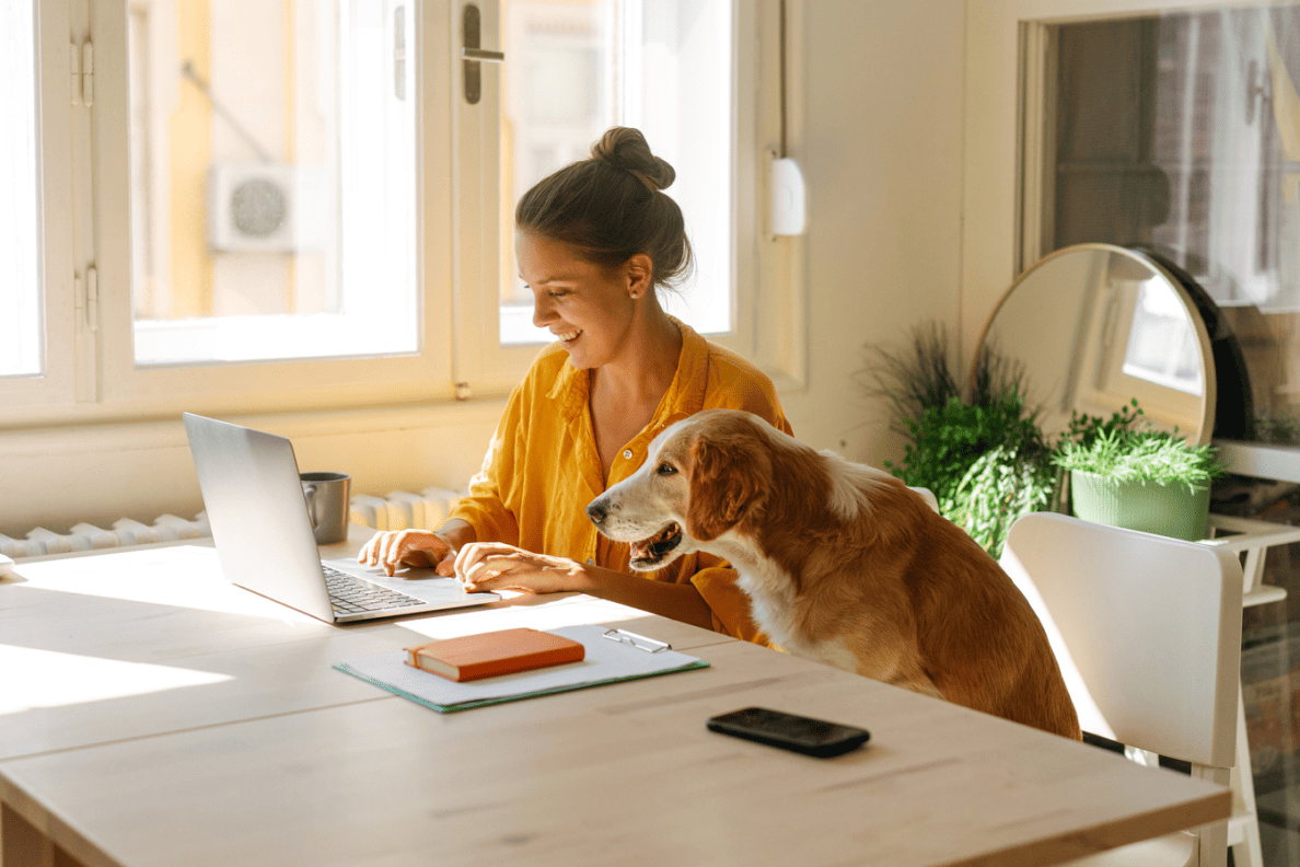 Une femme souriante attablée devant son ordinateur, un chien assis à ses côtés.