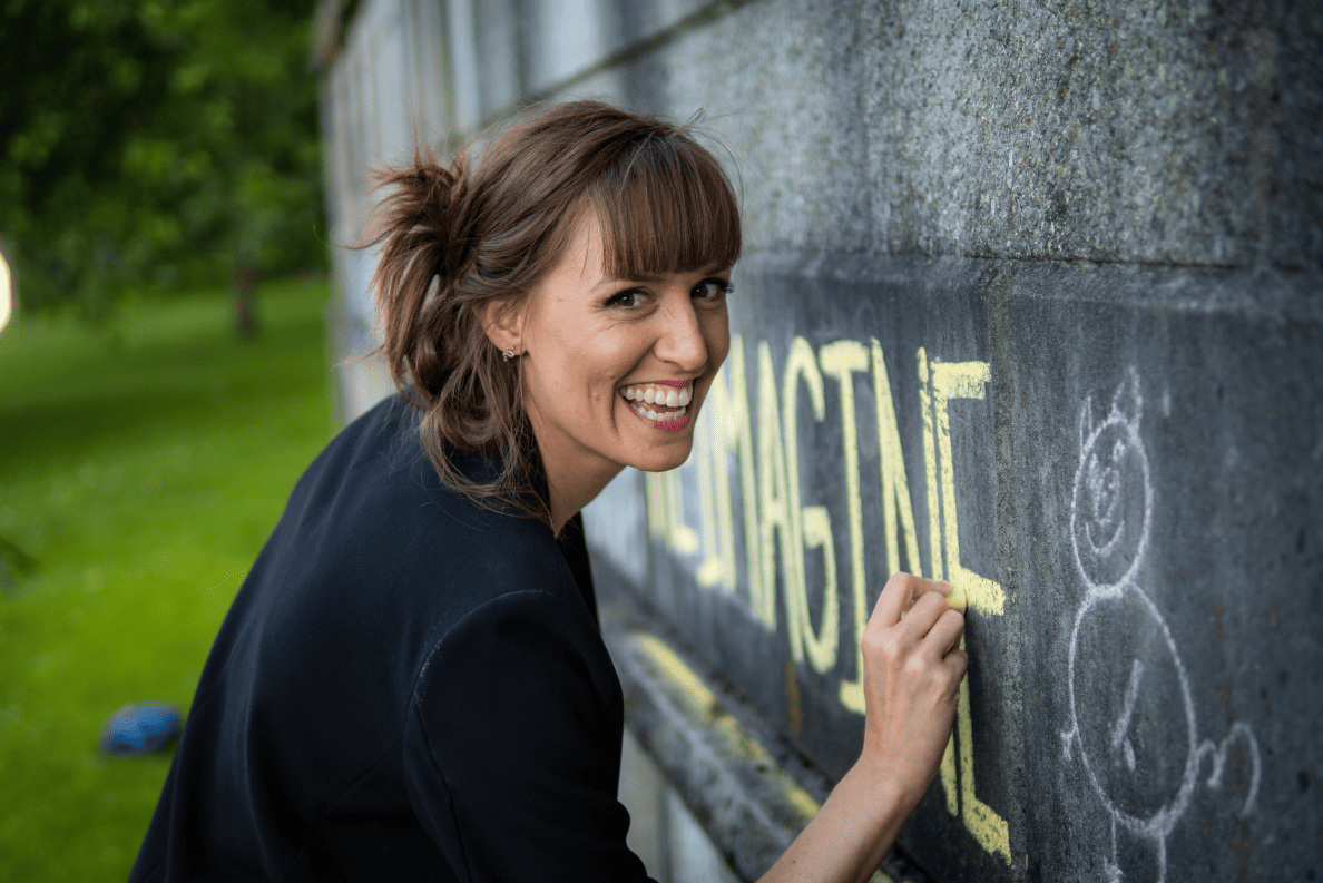 La futurologue Lindsay Angelo écrit à la craie jaun sur un mur de pierre noire le mot « imagine » en regardant la caméra en souriant à pleines dents.