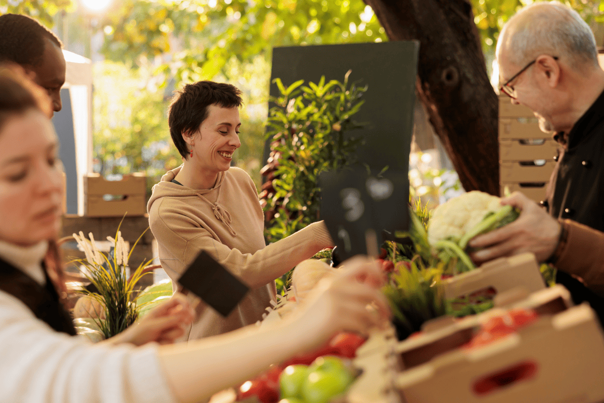 Une femme souriante choisit des légumes frais dans un marché extérieur, sous des arbres baignés de soleil.