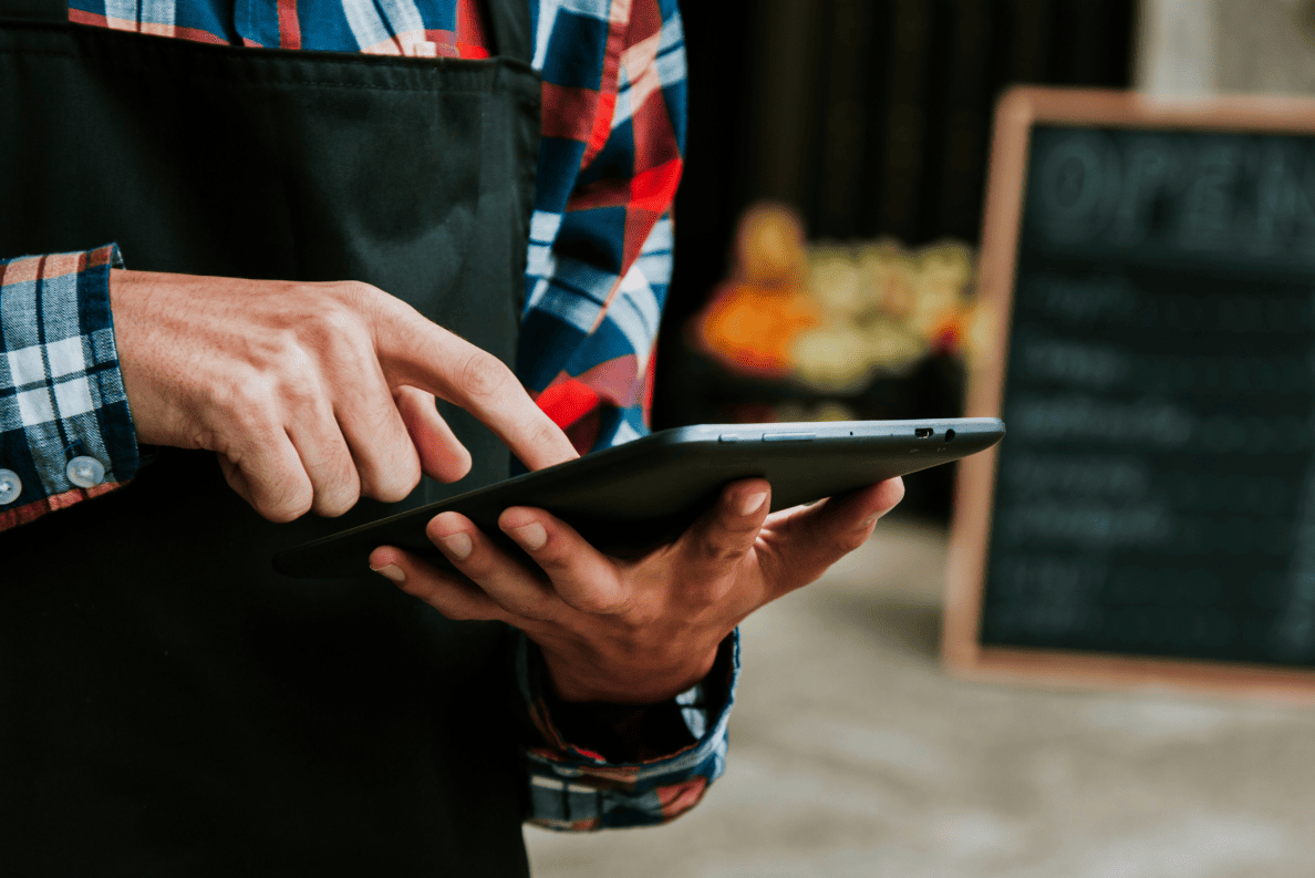 Un commerçant en tablier devant son étal de fruits et légumes manipule sa tablette intelligente.