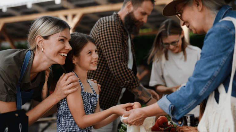 Le propriétaire d’un commerce présente ses produits à une femme et sa fille dans un marché extérieur animé.