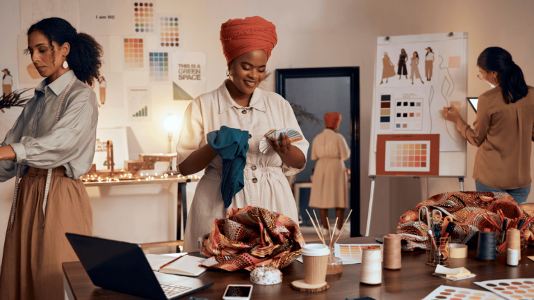 Trois femmes s’affairent dans un atelier de création rempli de tissus, croquis et échantillons.