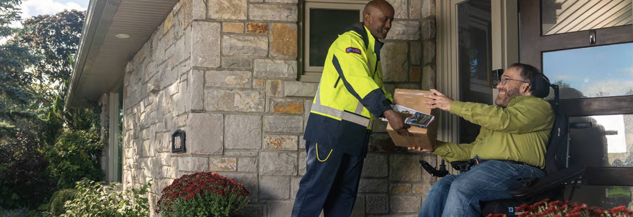 A Canada Post delivery agent hands a package to a man in a wheelchair at his front door. 