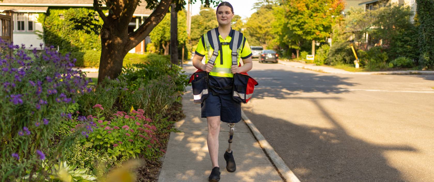 A mail carrier with a prosthetic leg carries mail delivery bags as she walks along a residential street. 