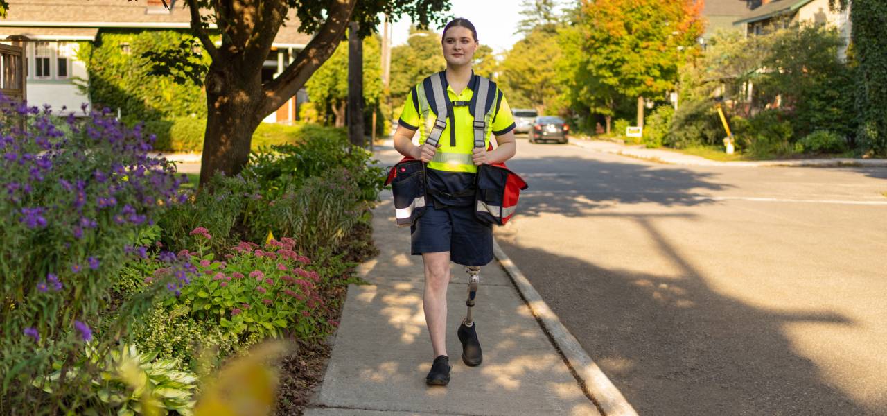 A mail carrier with a prosthetic leg carries mail delivery bags as she walks along a residential street. 