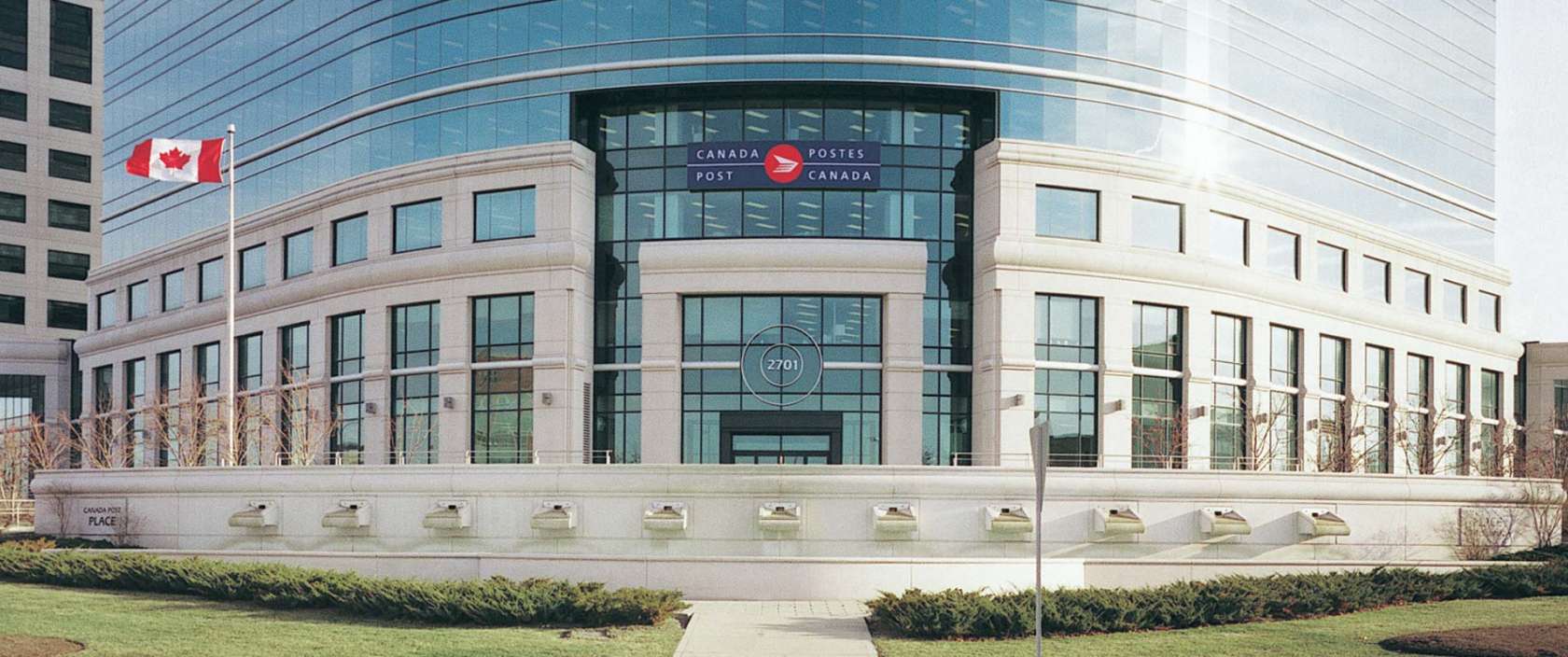 A Canadian flag flies in front of the Canada Post headquarters. The building features granite columns and expansive glass windows.