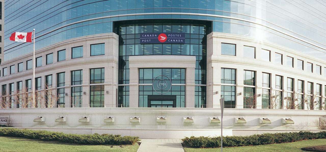 A Canadian flag flies in front of the Canada Post headquarters. The building features granite columns and expansive glass windows.