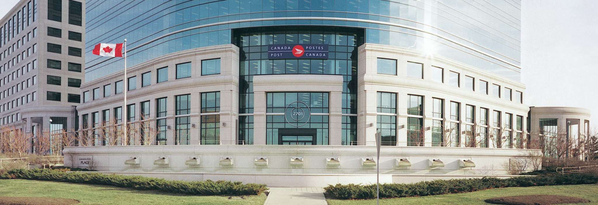 A Canadian flag flies in front of the Canada Post headquarters. The building features granite columns and expansive glass windows.