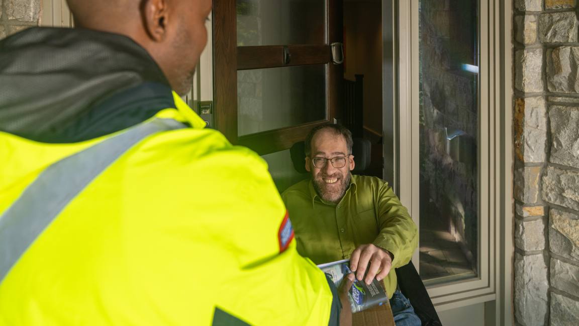 A man in a wheelchair accepts a delivery from a mail carrier at his front door.