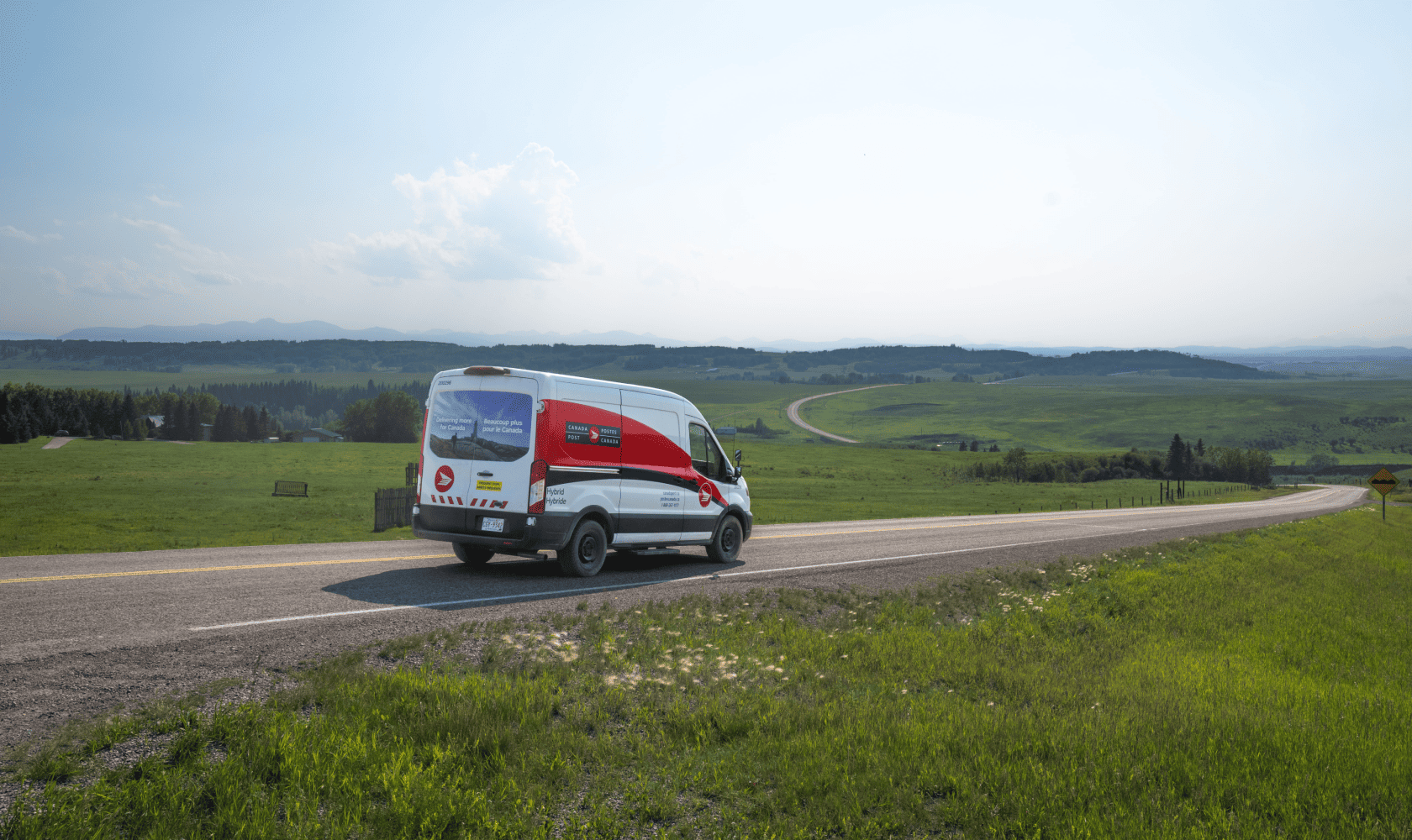 Canada Post delivery van driving along a rural highway through rolling green hills under a hazy sky.