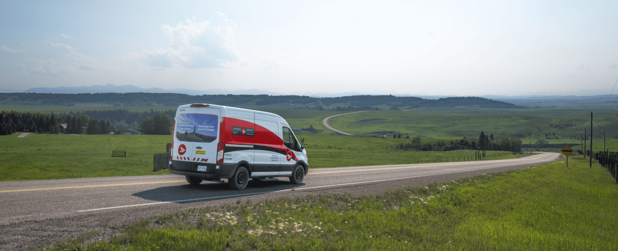 Canada Post delivery van driving along a rural highway through rolling green hills under a hazy sky.