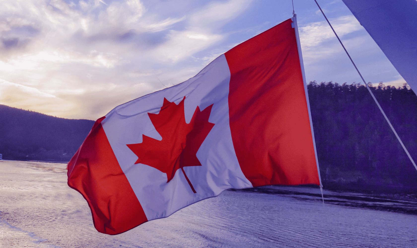 A Canadian flag waving with a body of water and forested hills in the background at sunset.